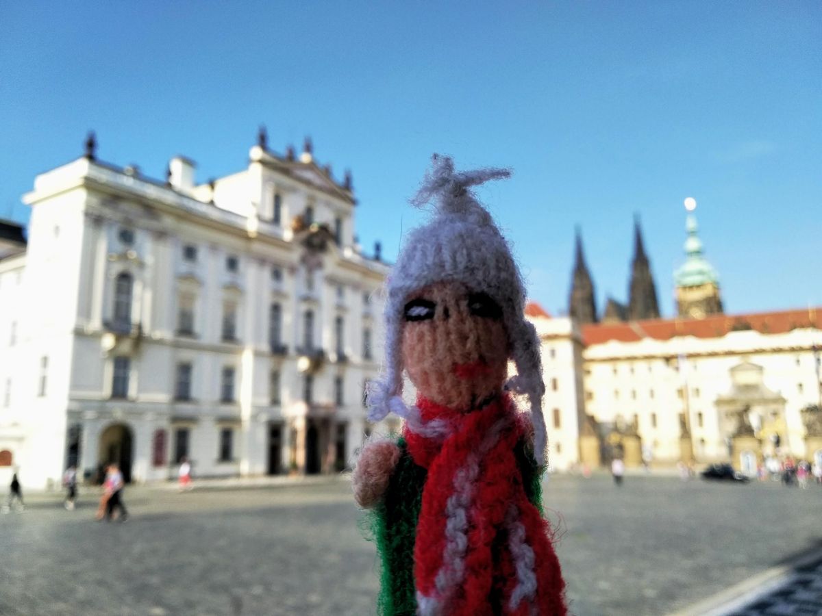 An erect and eager Peruvian finger puppet a white hat and red and white scarf, stands in the foreground with the blurred Prague Castle in the background.