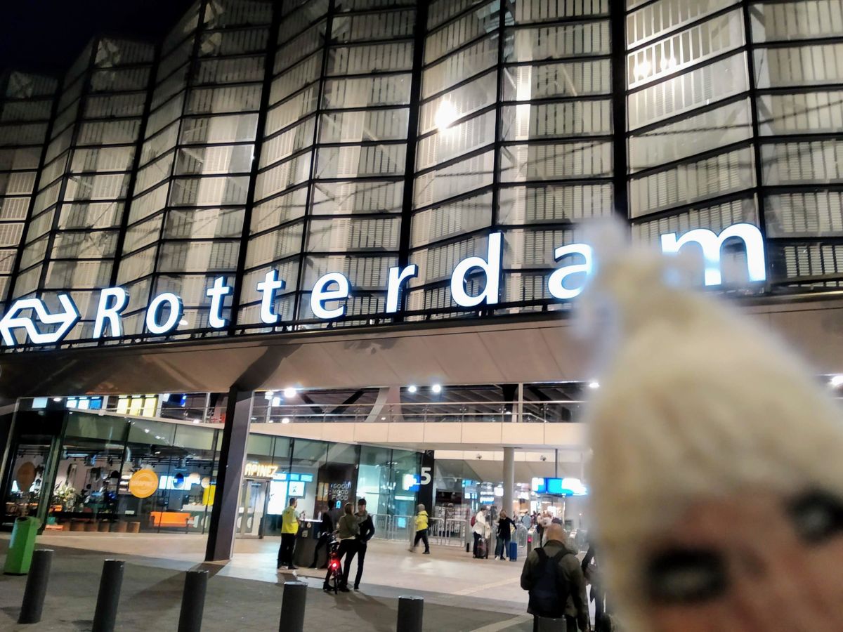 Nighttime exterior of Rotterdam Central Station's illuminated sign, with a finger puppet in the foreground partially obscuring the view.