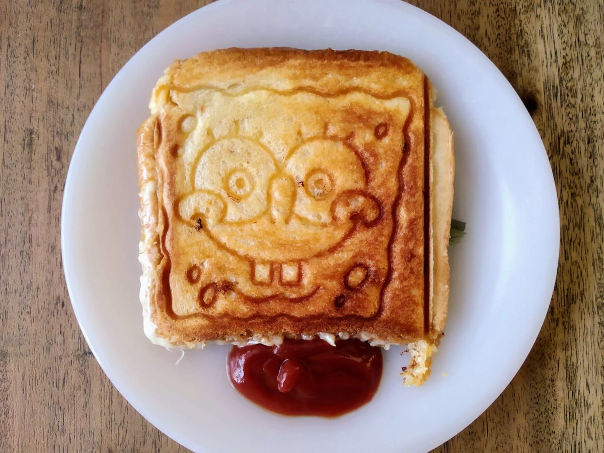 A grilled cheese sandwich with an embossed image of Spongebob Squarepants on the top slice, served on a white plate with a side of ketchup.
