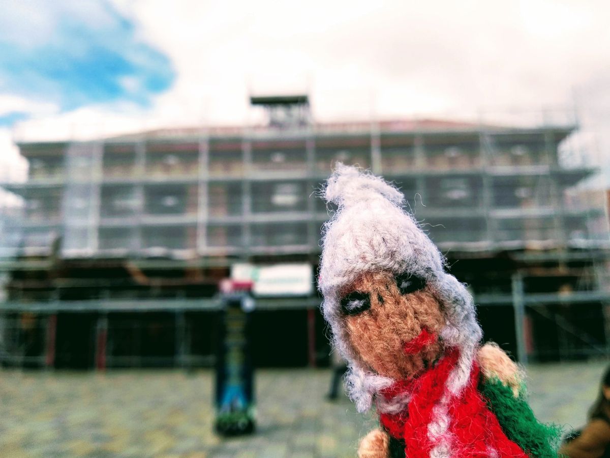 An industrious hand-knitted finger puppet in front of a scaffold-covered Saint-Étienne Châteaucreux.