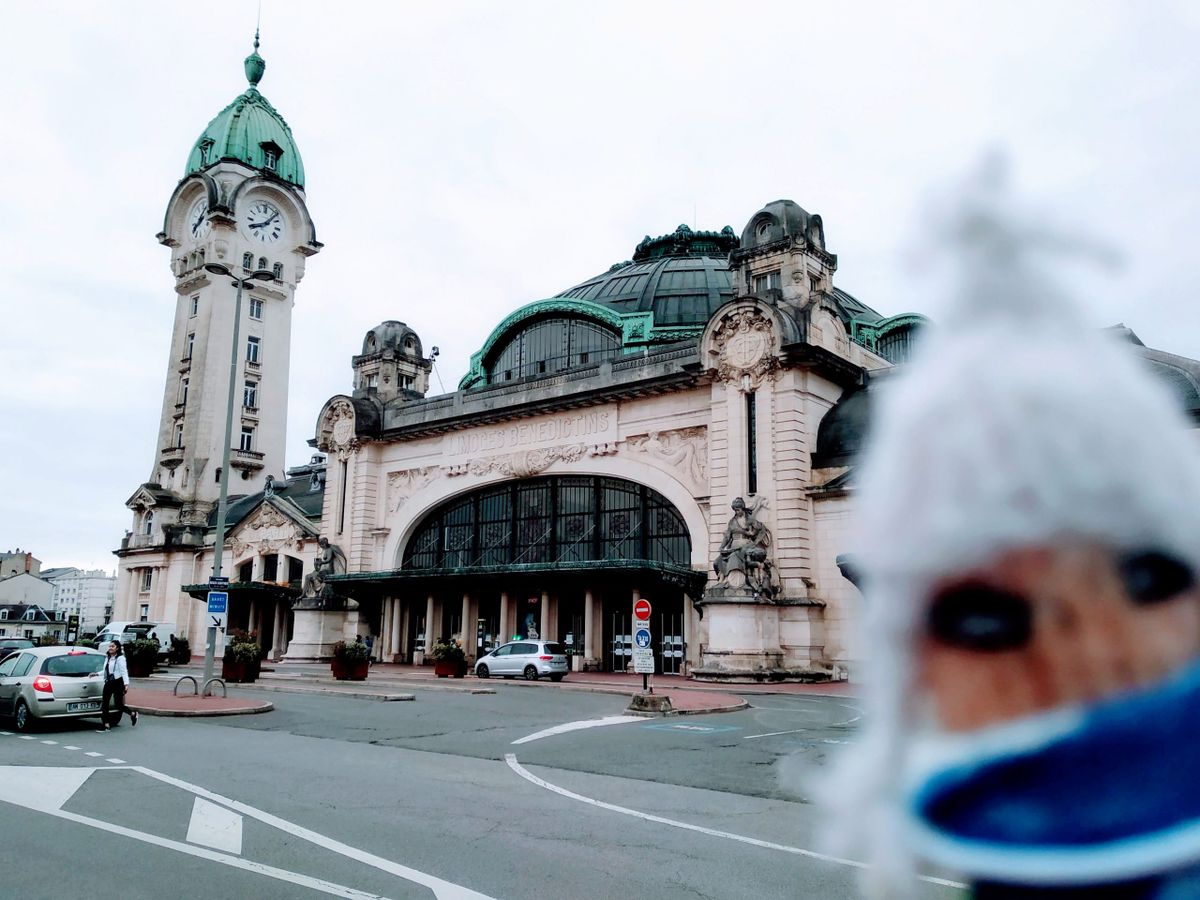 The ornate Limoges-Bénédictins train station, with a tall clock tower and green copper dome, is obscured by a finger puppet wearing a mask.