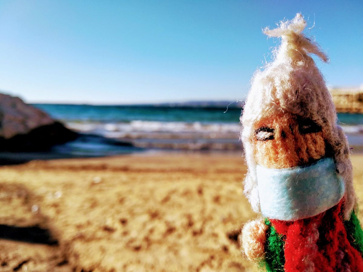 A wind-swept, sun-drenched finger puppet wearing a face mask on a sandy beach with the ocean and blue sky in the background.