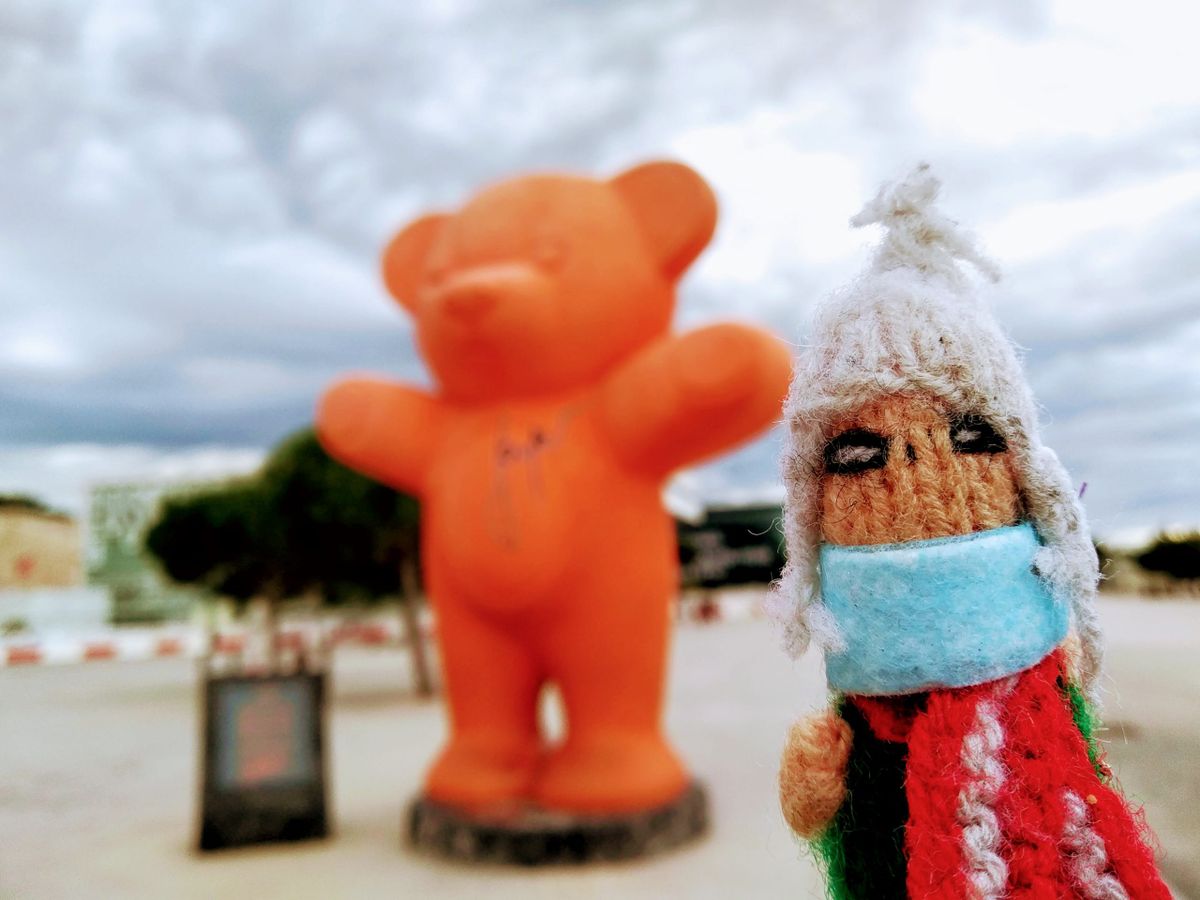 An art-loving finger puppet in a face-mask in front of a large orange bear statue in the blurry background.