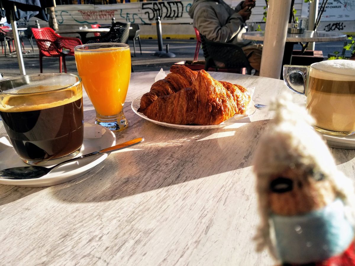 A sunny outdoor café table with a croissant, a glass of orange juice, a coffee, and a latte. A small finger puppet in a face mask is visible in the foreground.