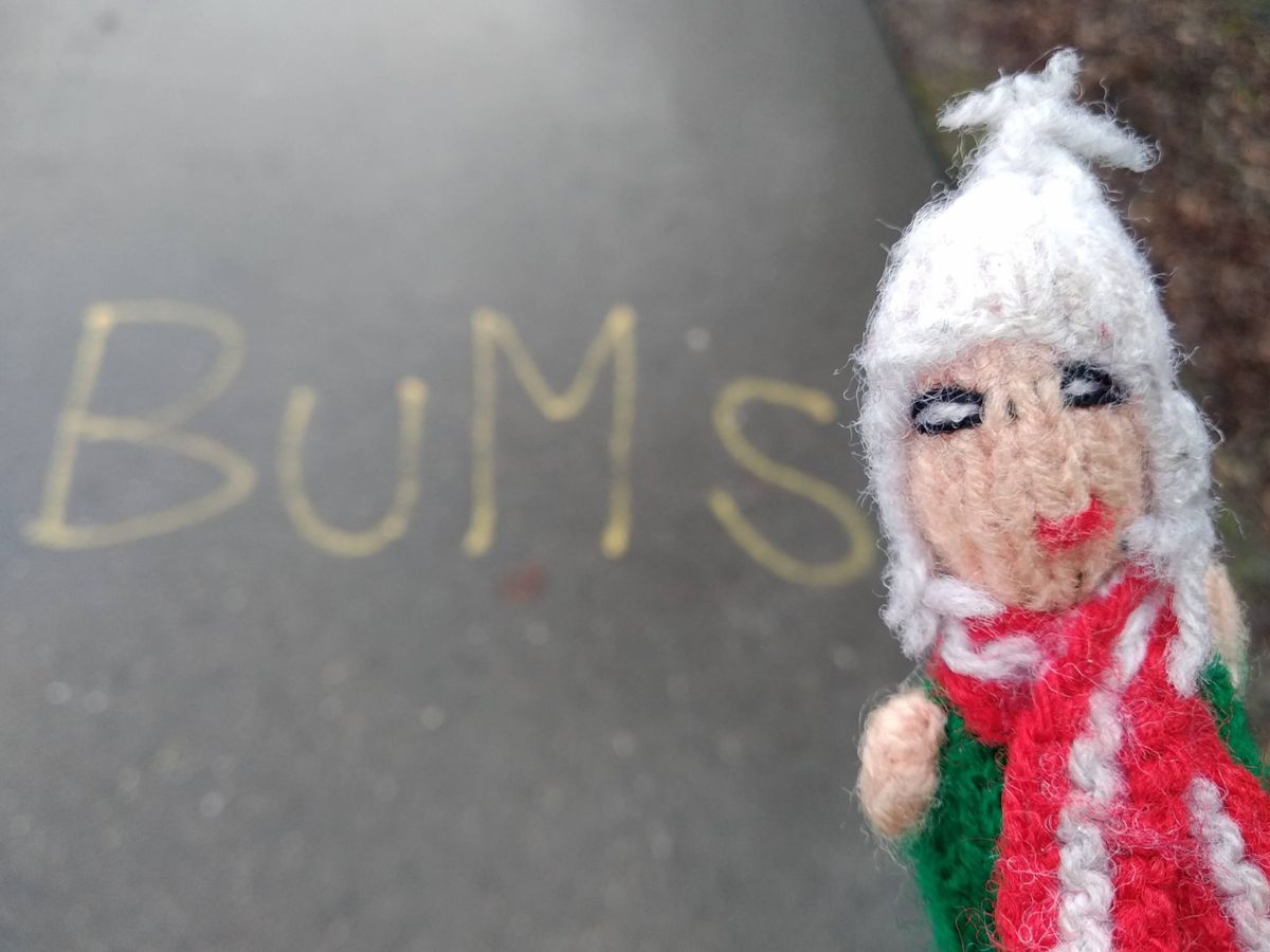An easily-amused finger puppet in front of the word "BUMS" written in chalk on a road.