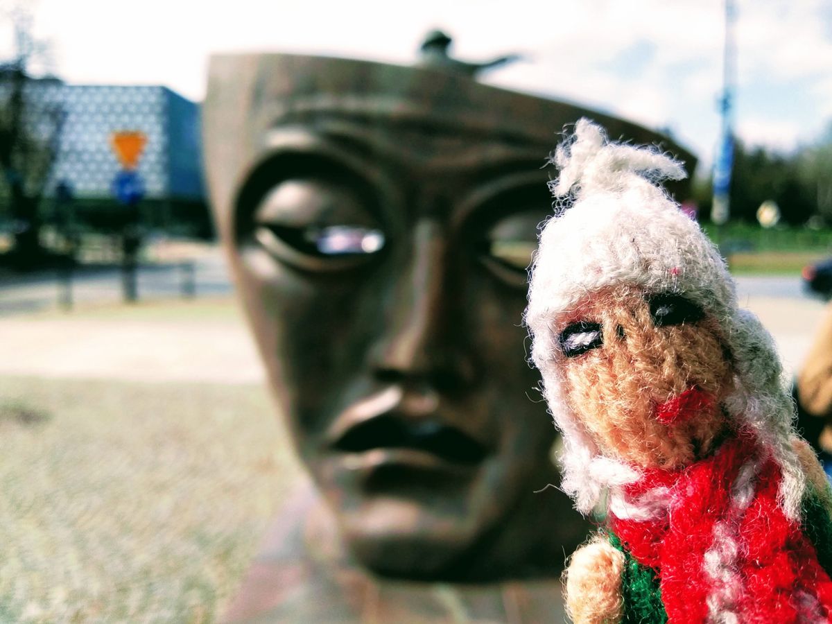 A tired finger puppet in front of a bronze tragedy mask on a pavement.