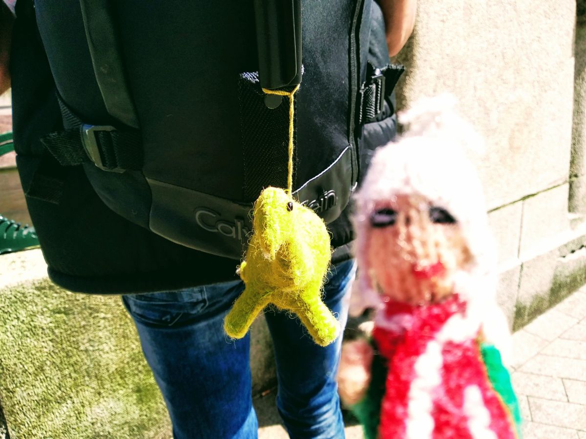 A coquettish Peruvian finger puppet turns away in shame from a cheeky yellow dinosaur hanging from a confused man's backpack.
