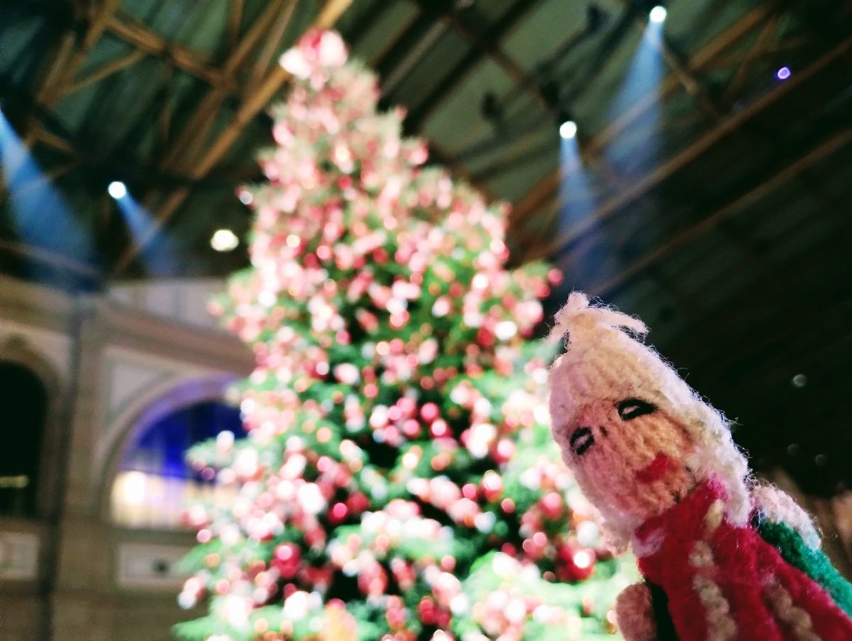 A festive Peruvian finger puppet tries to blend into a massive Christmas tree decorated in green, red, and white lights.