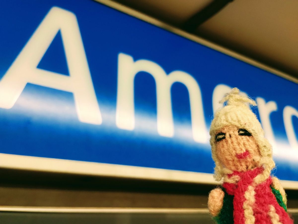 A finger puppet in front of the blue station sign at Donostia Amara Euskotren station.