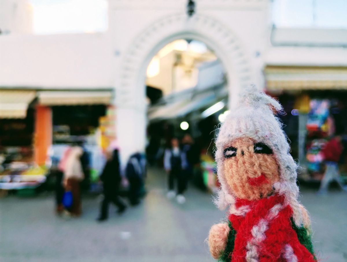 Puppet in front, bustling market street and the large white archway of Tangier's Bab Rahbat.