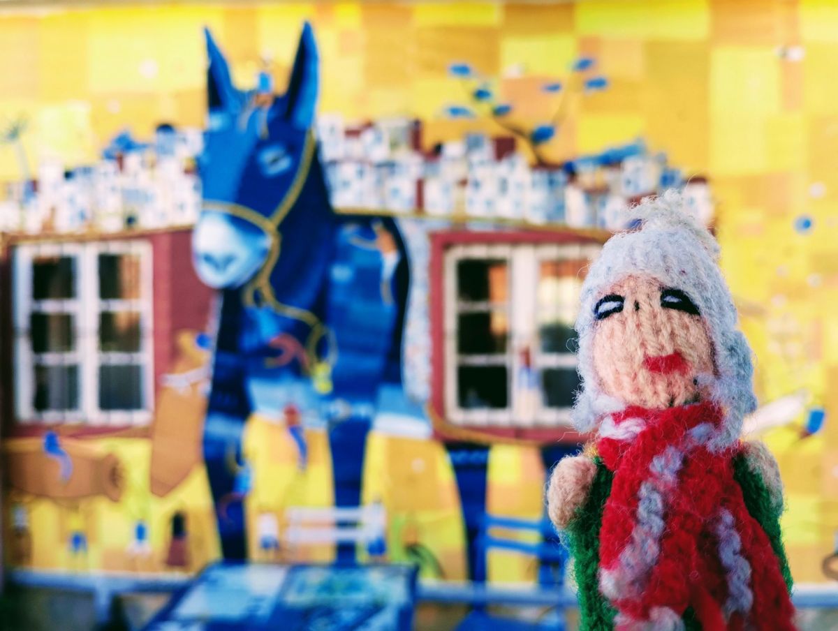 A stout yet strong Peruvian finger puppet in front of a painted mural of a blue donkey carrying a city on its back, against a yellow wall..