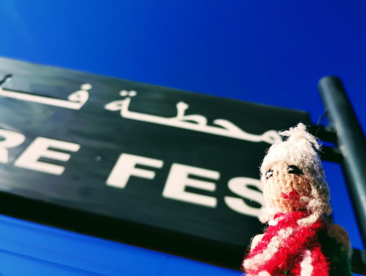 Against a blue sky, a mercenary Peruvian finger puppet inspects a platform sign in Arabic and English at Gare Fès.