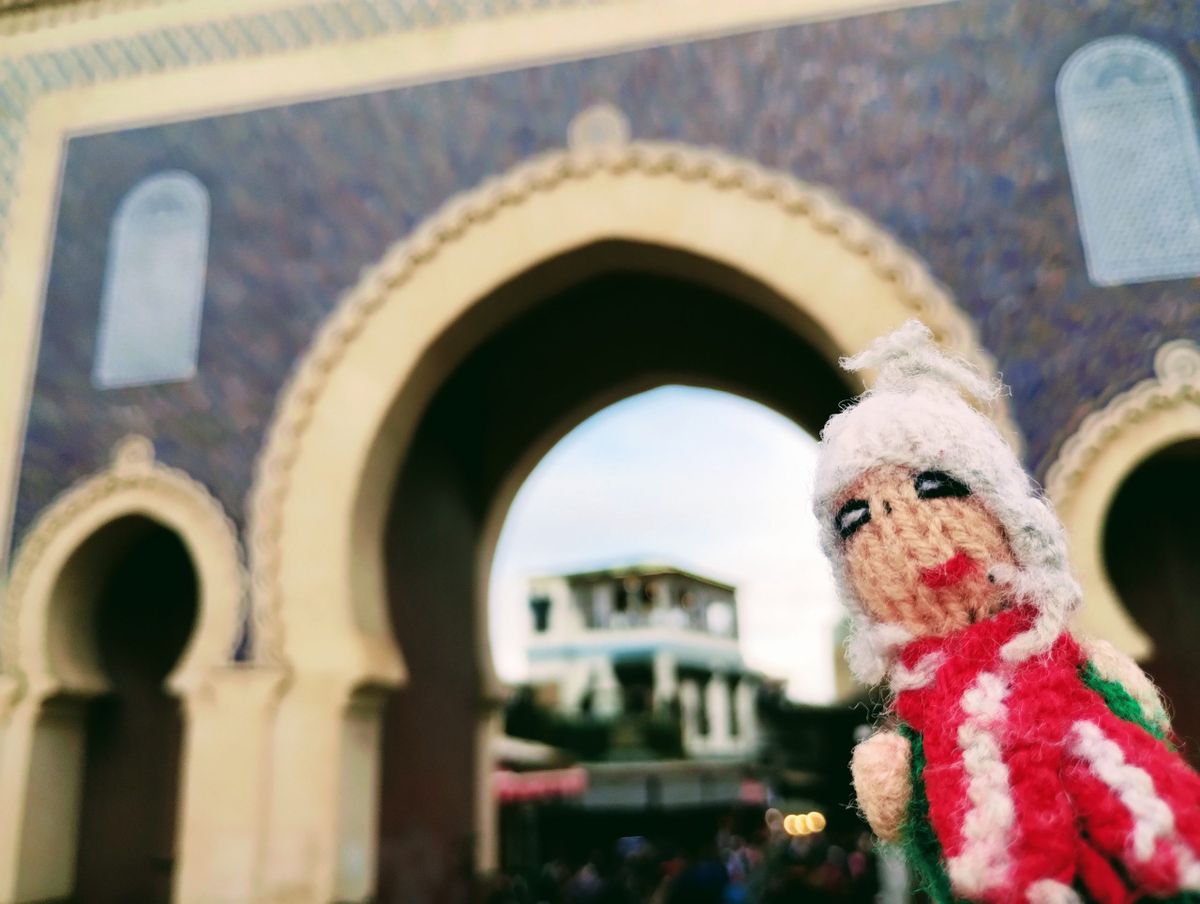 Finger puppet in front of the traditional Moroccan horseshoe archway of the Bab Boujloud in Fès, with blue and gold mosaic tiles and buildings visible through the horseshoe arch.