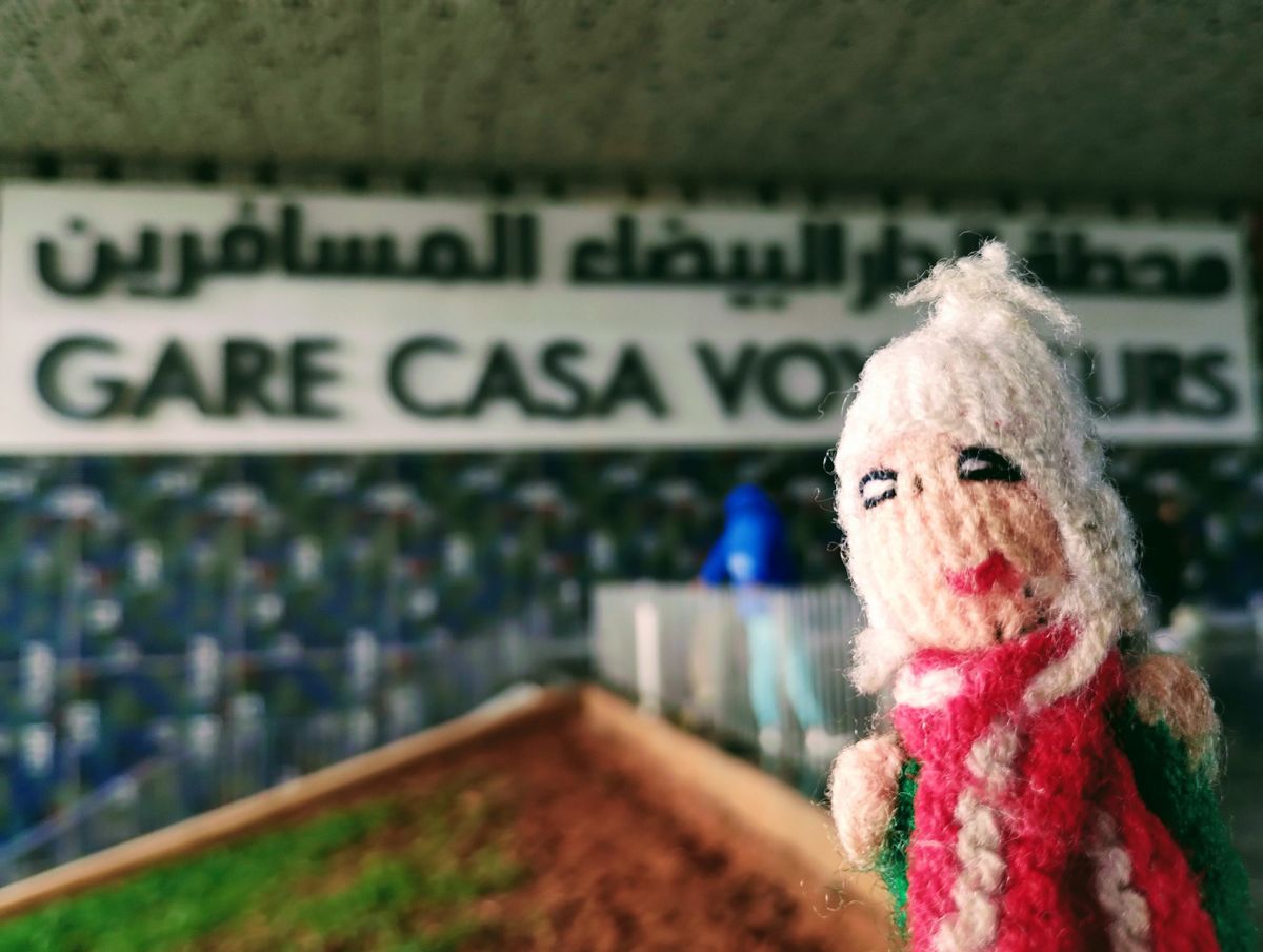 A glorious finger puppet standing in front of the "Gare Casa Voyageurs" train station sign.