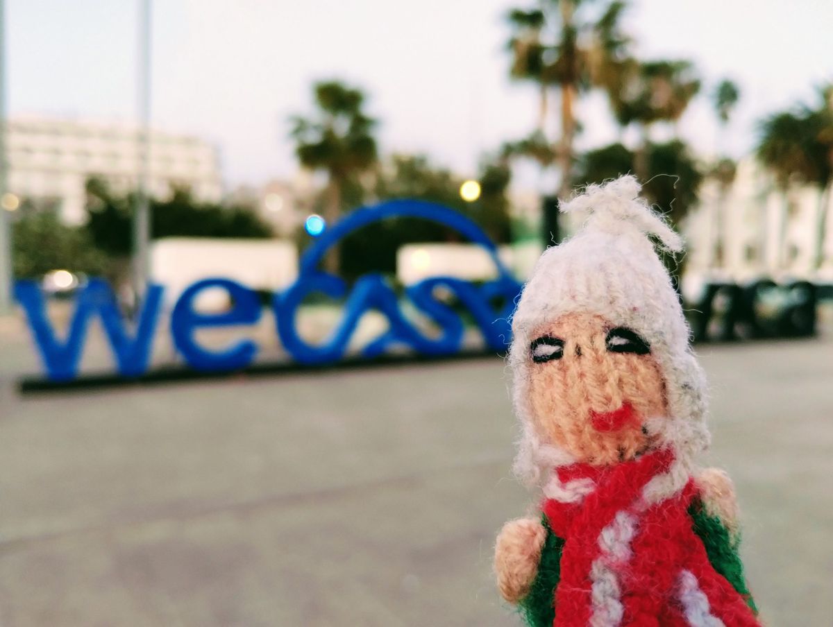 A small Peruvian finger puppet, standing in front of large blue letters of the "WeCasa" sign. In the background, palm trees and buildings against a slightly overcast sky.