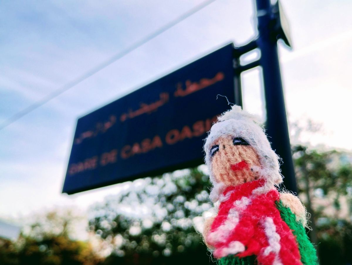 A bristling finger puppet positioned in front of a dark blue sign that reads Gare de Casa Oasis. The background shows a blue sky and green foliage.