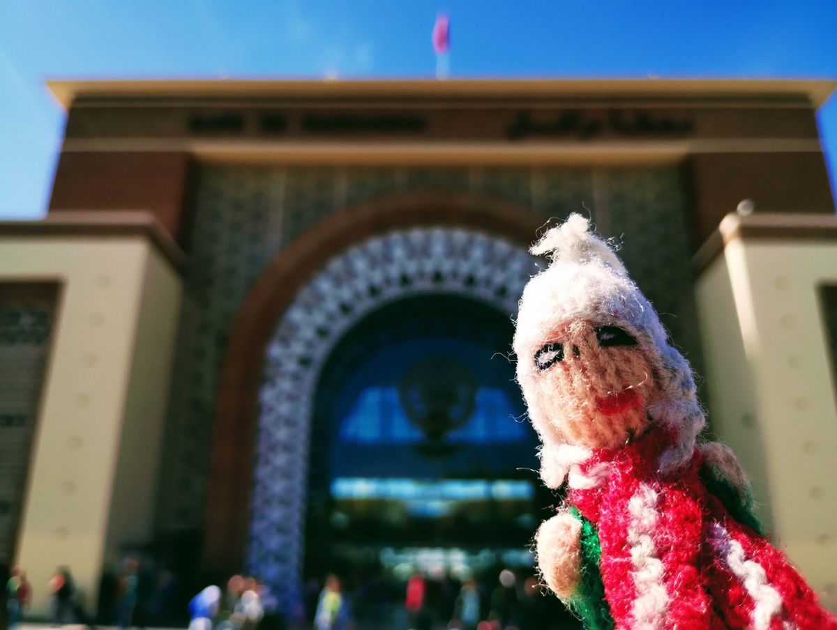 A well-travelled finger puppet in front of the large, ornate building arch of the entrance to Marrakesh train station.
