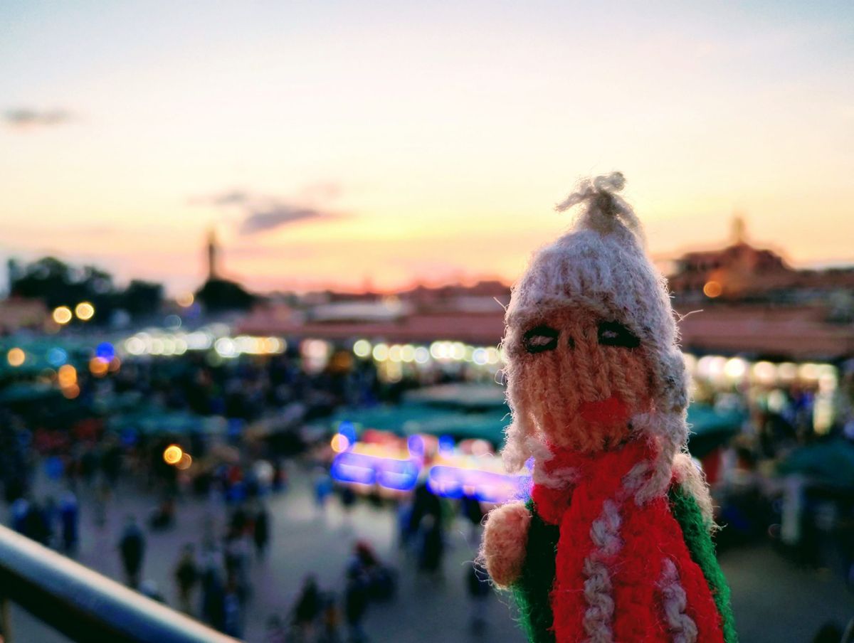 An aroused finger puppet in front of the Place Jemaa el Fna at sunset. The sunset is a gradient of oranges and pinks, and the city lights are bokeh and bright.