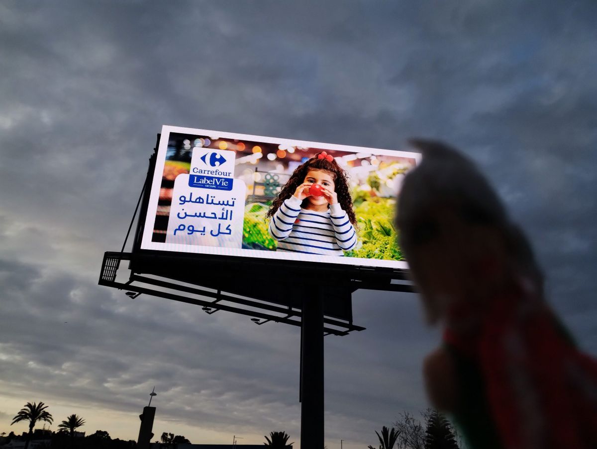 Capitalist finger puppet in front of a large Carrefour billboard showing a smiling girl. The Arabic text translates to 'You deserve the best every day.'