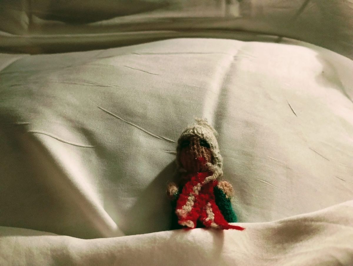 An exhausted Peruvian finger puppet lying against a pillow with a sheet pulled up over him.