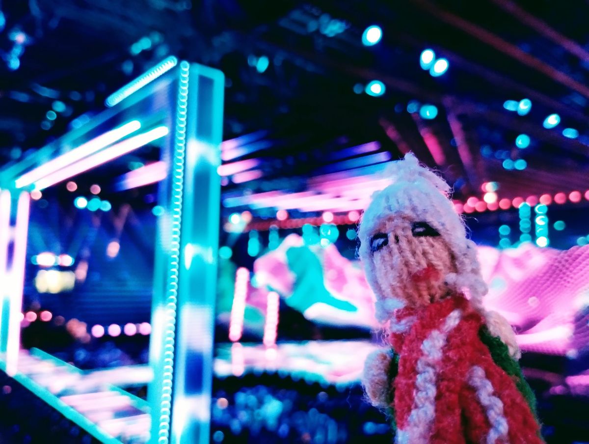 Finger puppet in front of the blurred blue and purple stage at the Eurovision Song Contest.