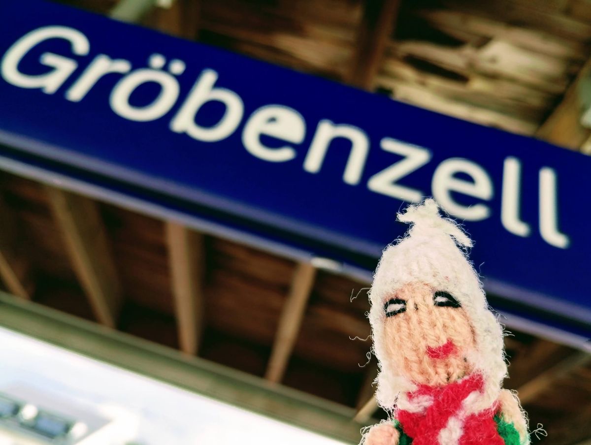 A happy puppet wearing a white hat and red scarf beneath a blue sign that reads "Gröbenzell".