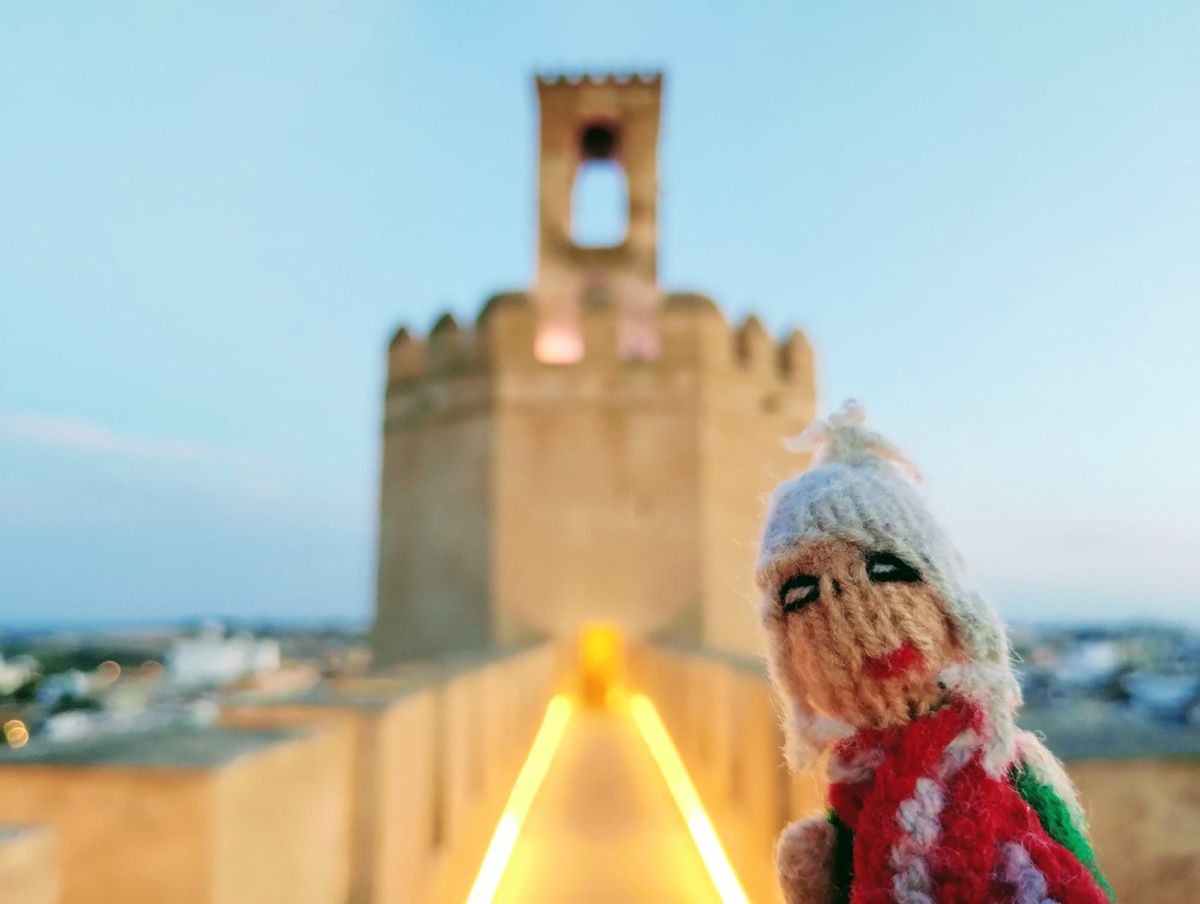 A magnificent finger puppet posing in front of a medieval fortress at dusk. A yellow light illuminates the path in front of the fortress.