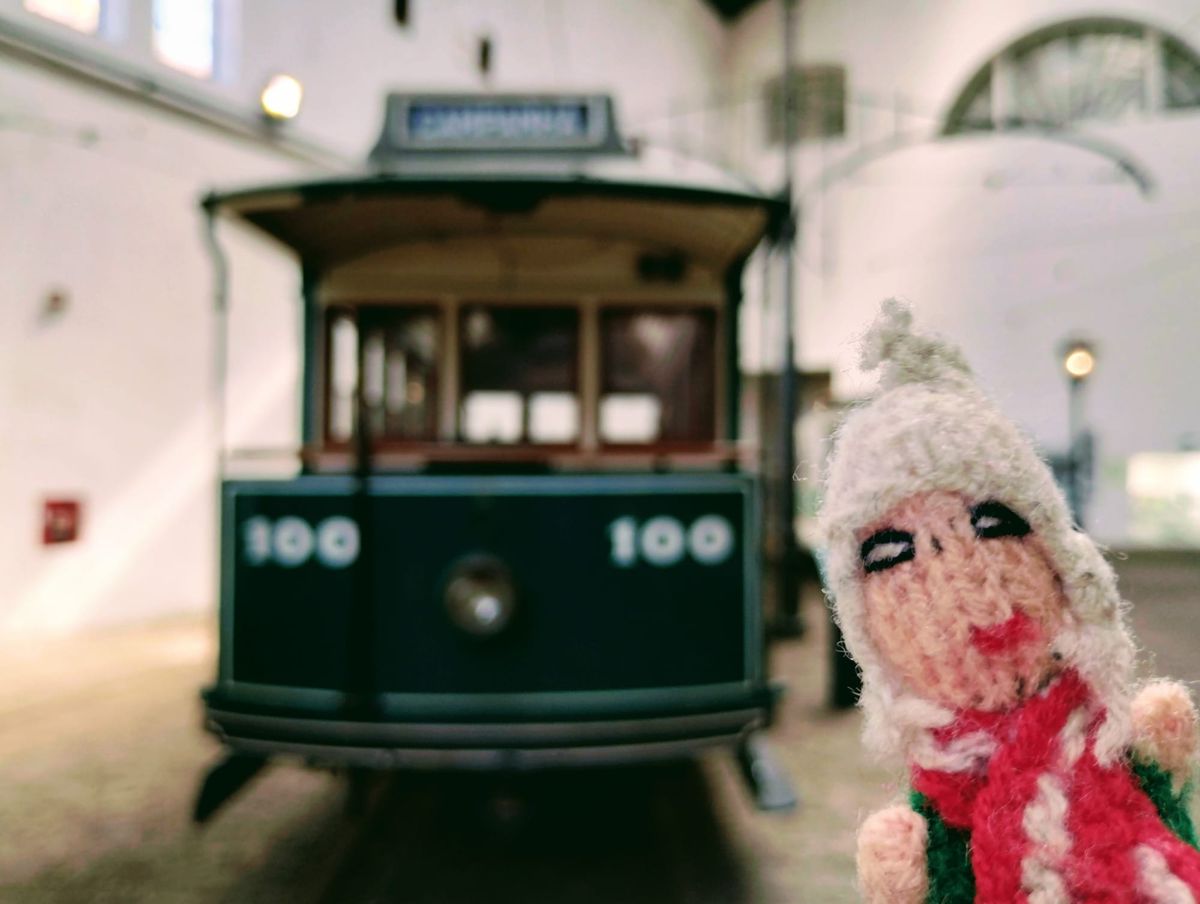 Finger puppet stands in front of a vintage dark green trolley car displaying the number 100, inside a building with white walls and architectural details.
