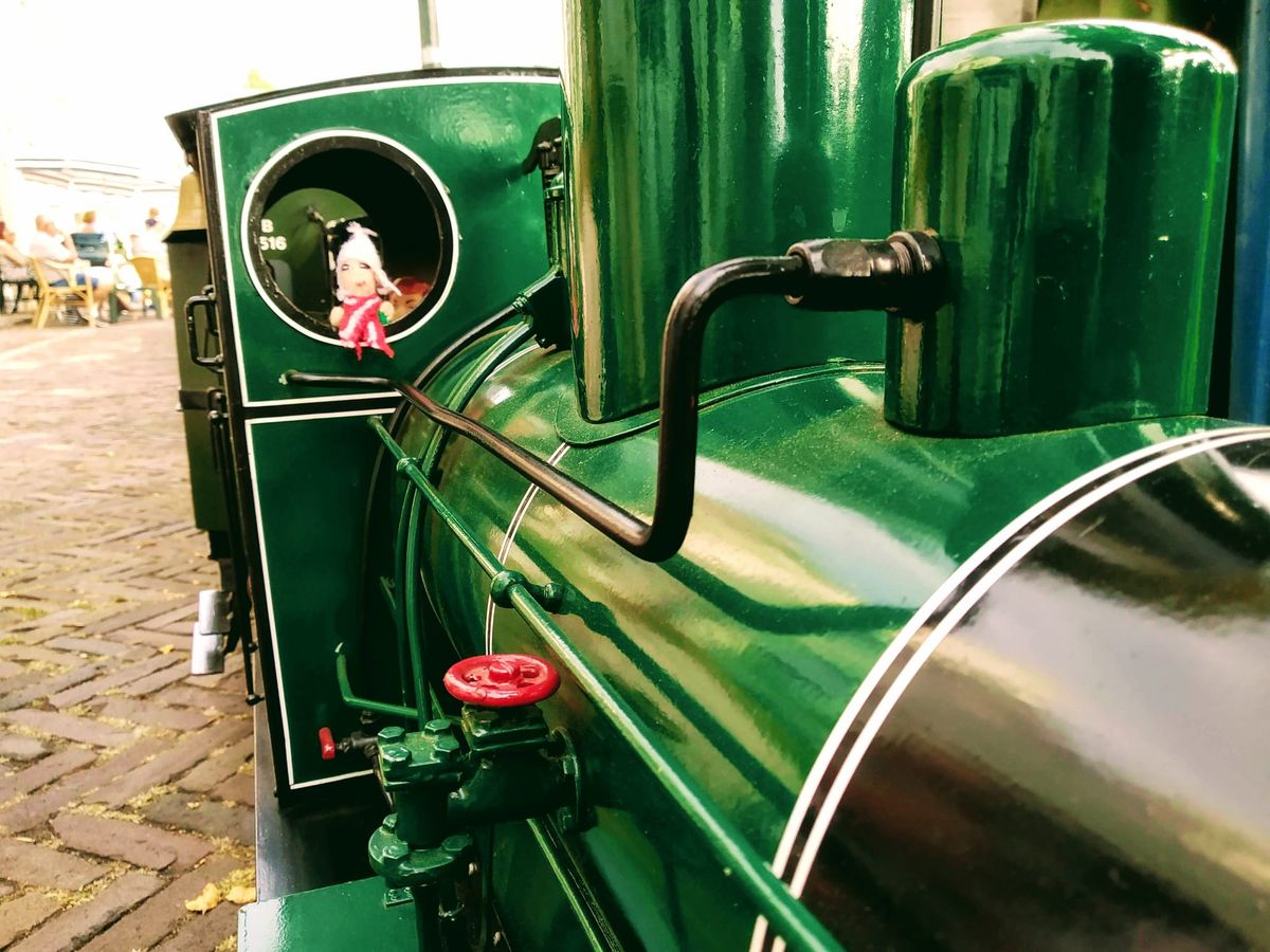 An enthusiastic finger puppet driving a detailed, green-painted miniature steam engine with various mechanical parts, including pipes, a red valve, and a circular viewing window.