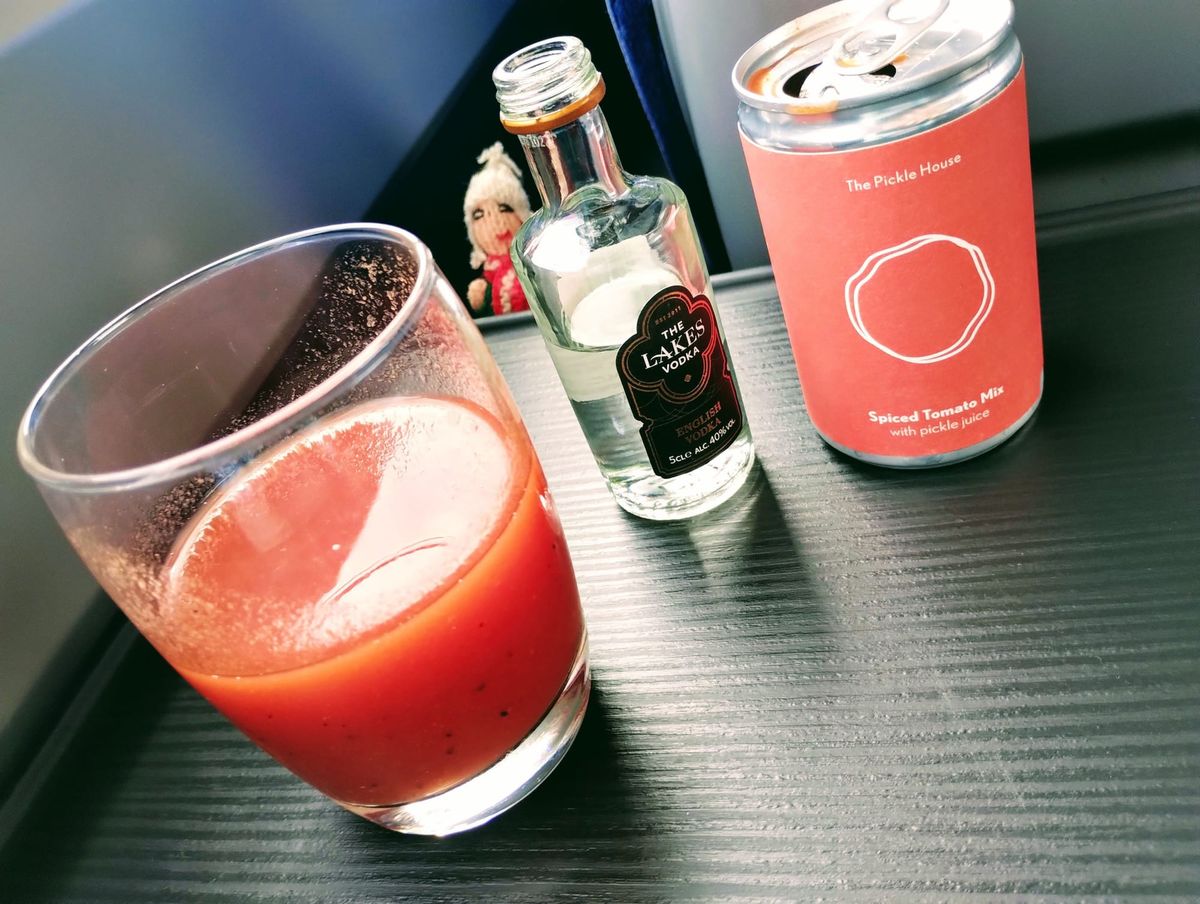 Finger puppet reels behind a glass of Bloody Mary, a mini bottle of vodka, and a can of spiced tomato juice are arranged on a train table.