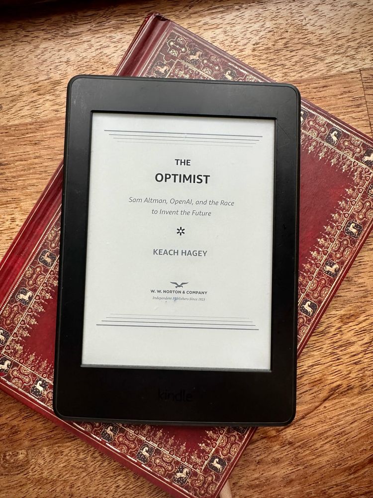 En Kindle ligger oppå en notatbok. På Kindle-skjermen vises coveret av boken The Optimist.
