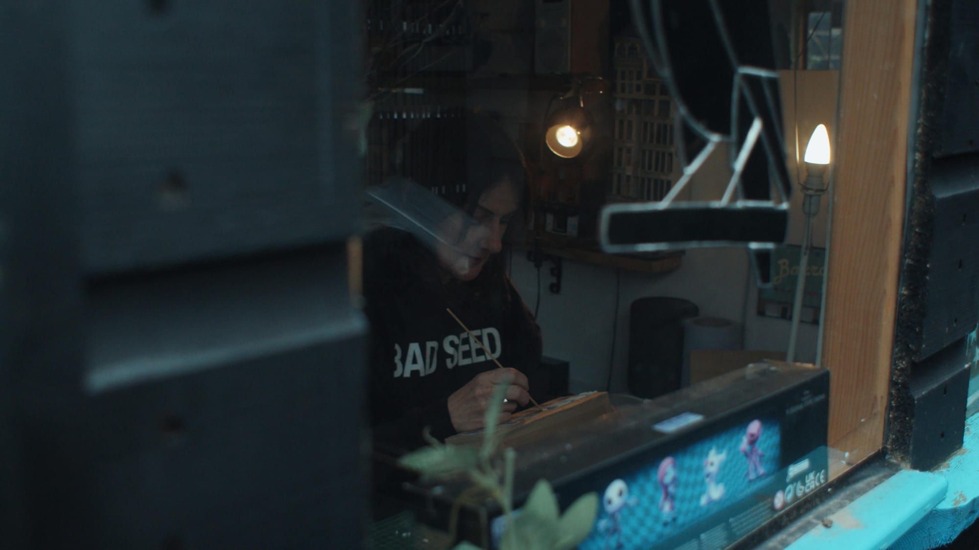 A woman painting a miniature inside a cosy art shed, seen through a window
