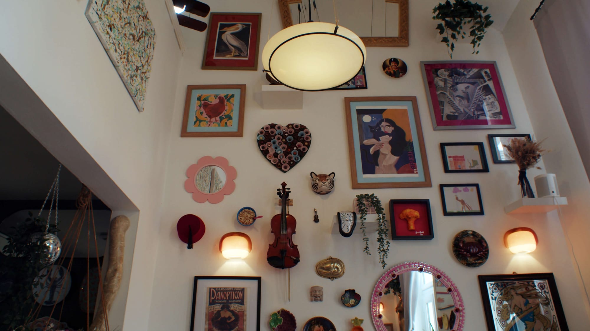 A duplex flat wall filled with eclectic art, mirrors, plants, and objects, including a violin, framed prints, masks, and a heart-shaped decoration