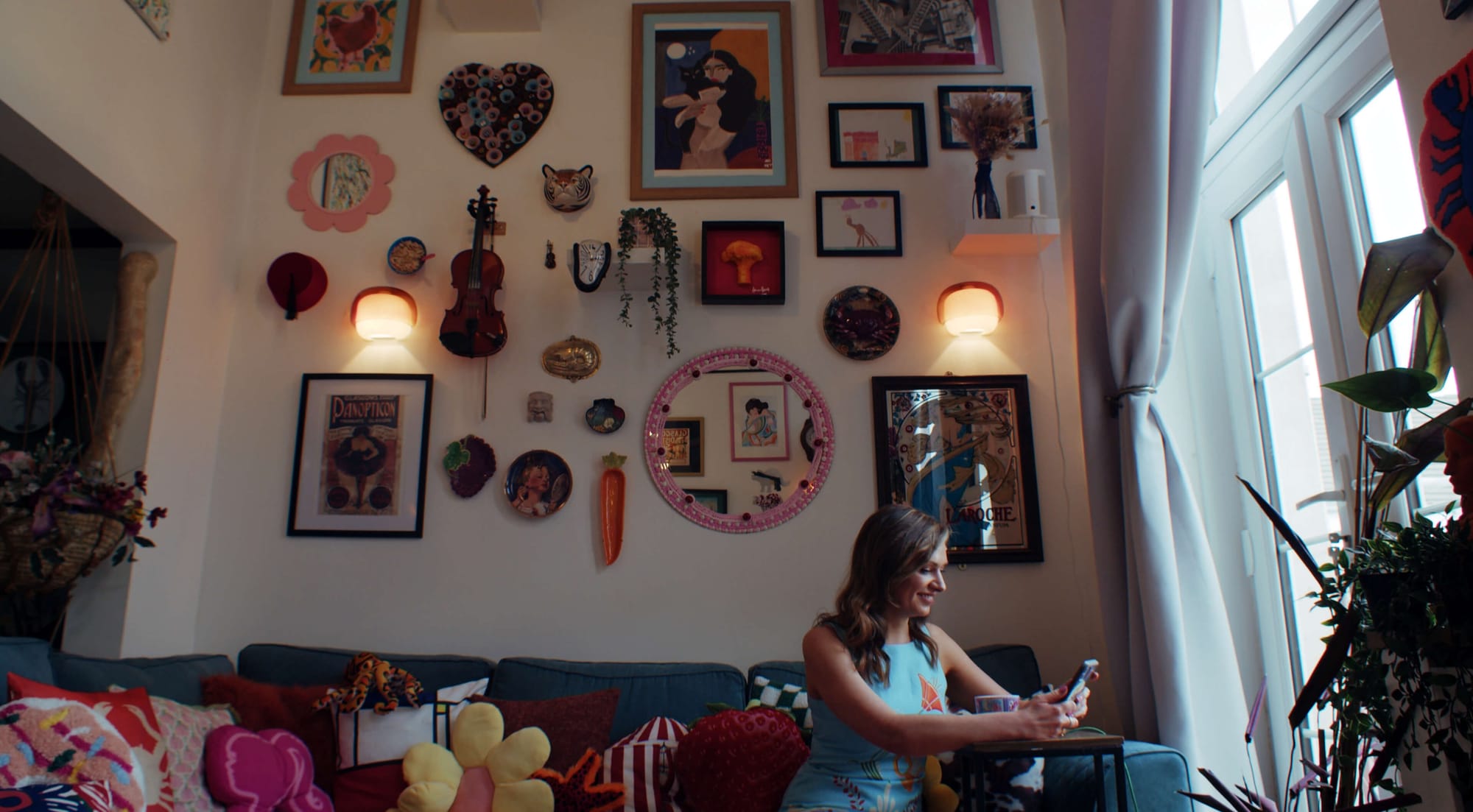 A bright living room with a colourful gallery wall and a woman in a blue dress sitting on the sofa