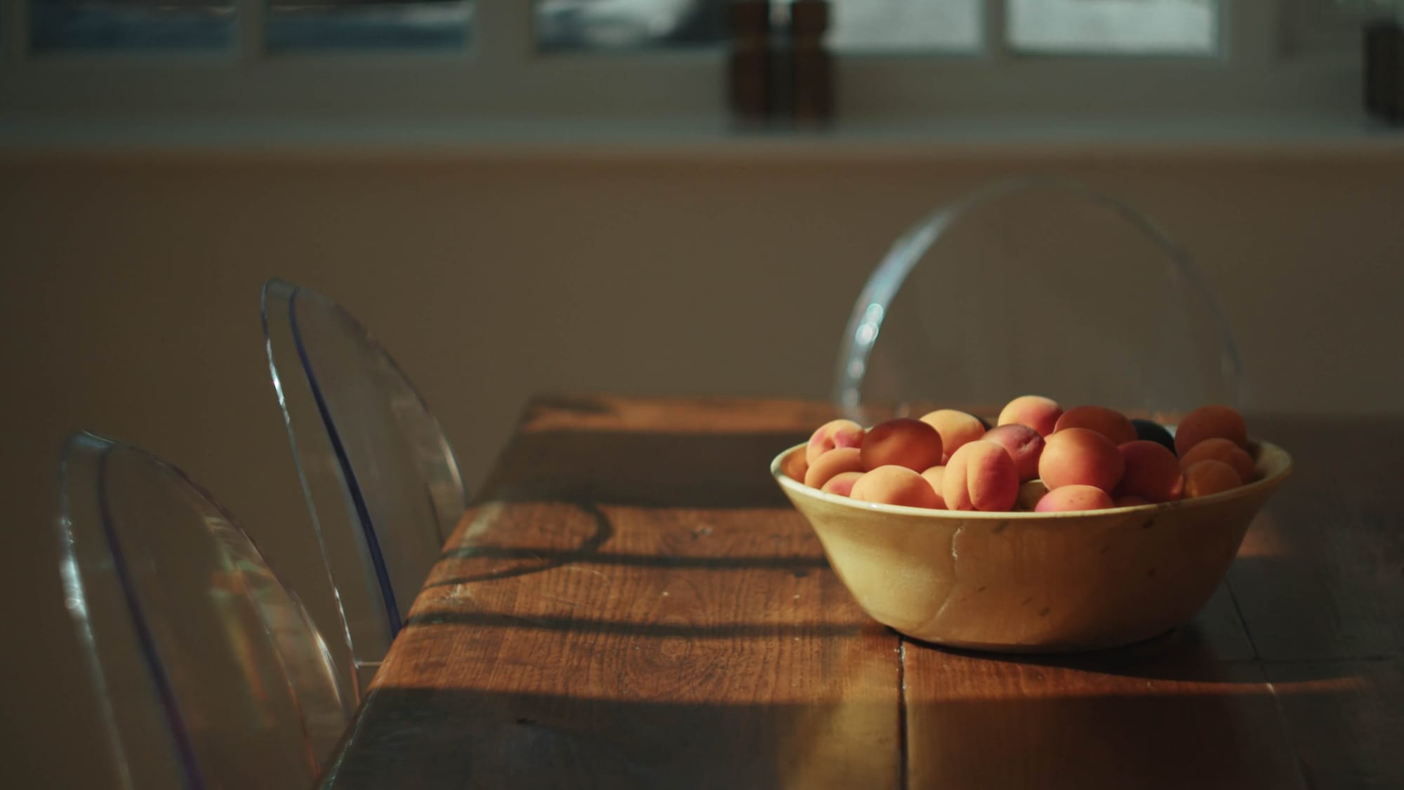 A sunlit wooden table with a bowl of ripe apricots and three clear ghost chairs
