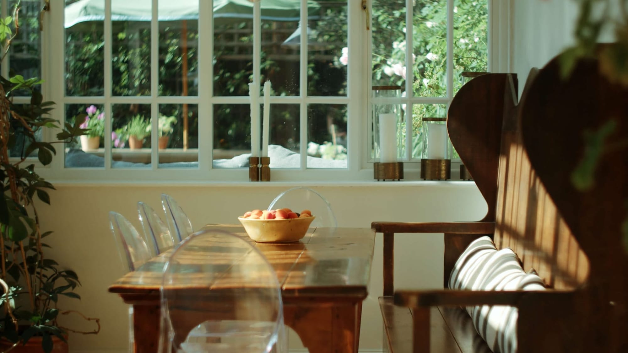 A bright kitchen nook with a wooden table, ghost chairs, and a settle bench facing a sunlit garden