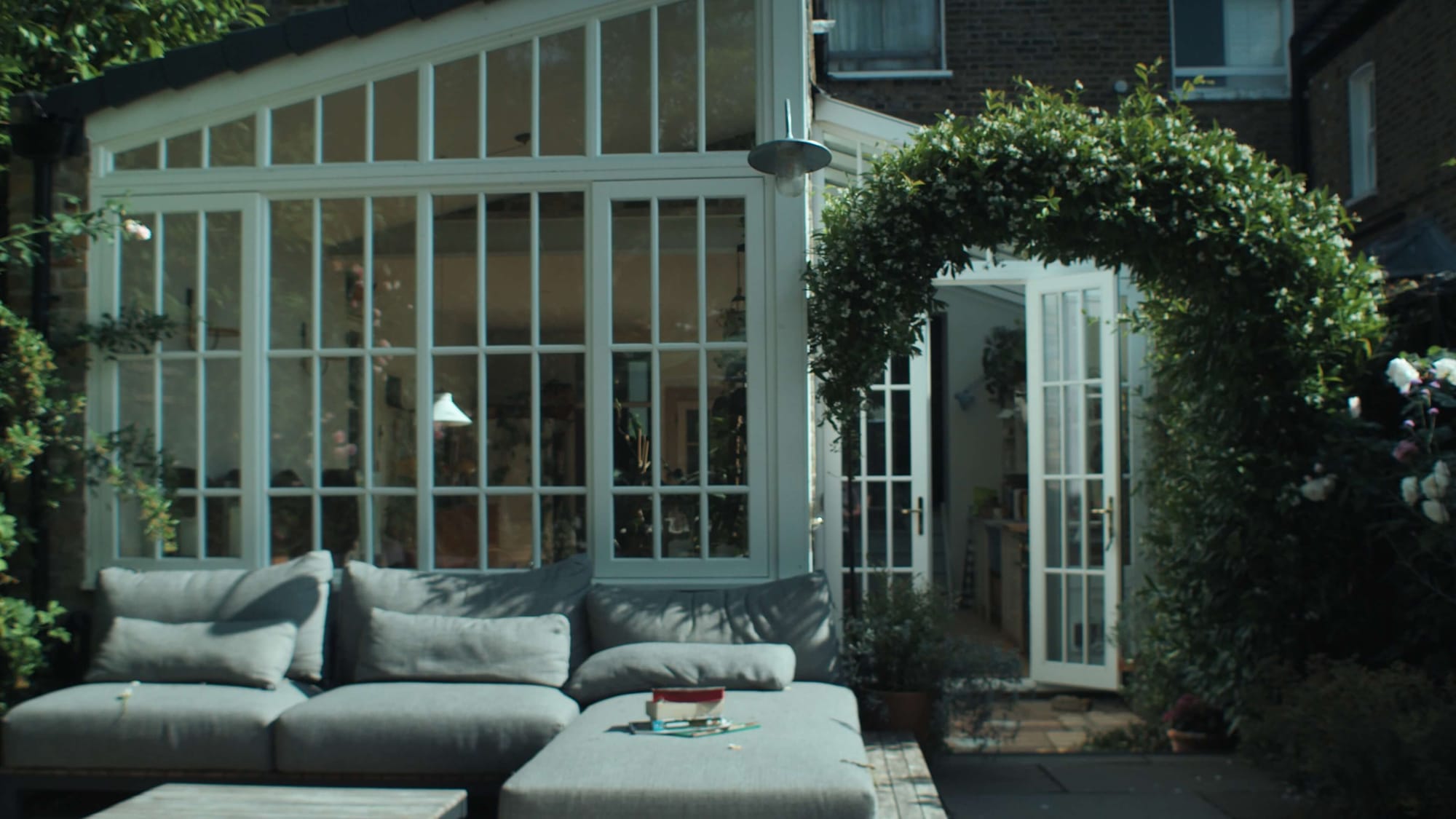 A leafy garden terrace with a grey outdoor sofa and a view into the glass-fronted kitchen