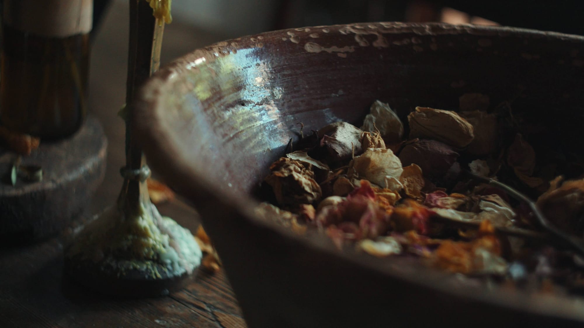 A rustic ceramic bowl filled with dried petals and wilted flowers on a wooden table, next to a candle holder thick with melted wax