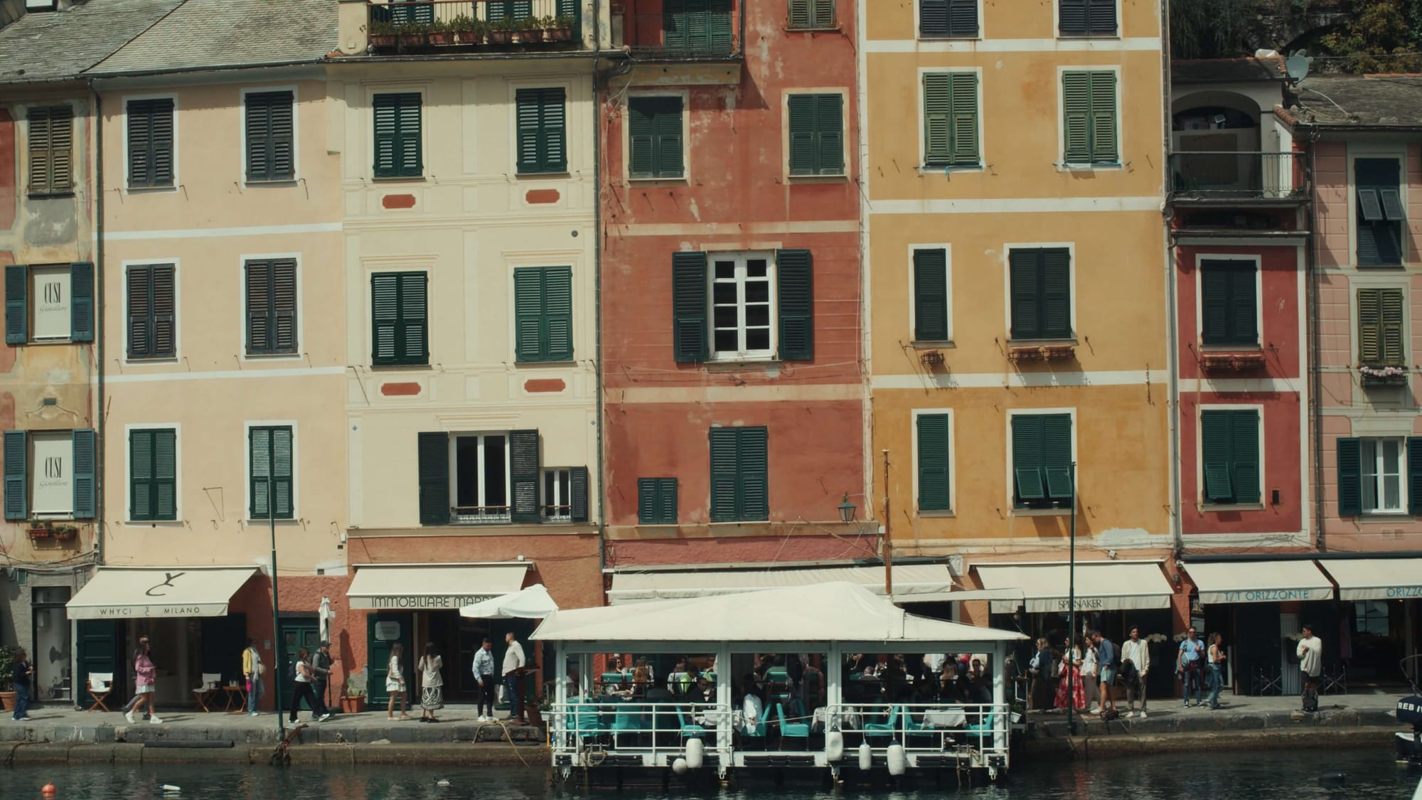 Colourful Ligurian waterfront buildings with faded pastel facades and green shutters, overlooking a busy promenade and floating café