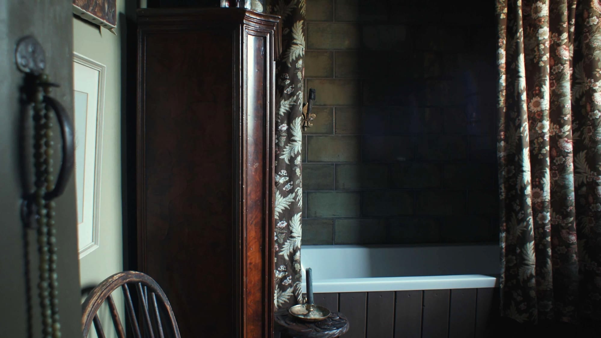 A dim, atmospheric bathroom with a white bathtub, dark tiled walls, and heavy floral curtains. A tall wooden cabinet stands beside a rustic chair, and a small carved table holds a candle and brass dish