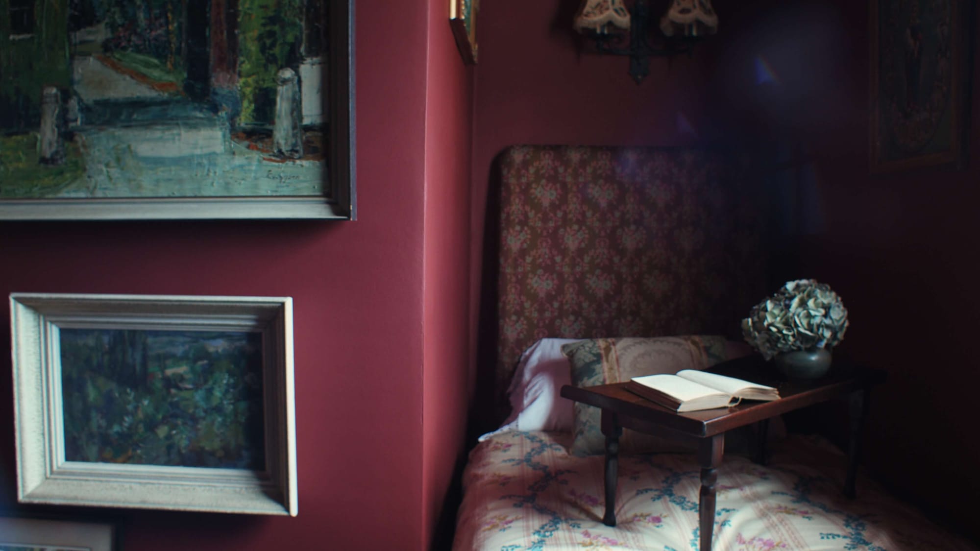 A small red bedroom nook with vintage paintings, a patterned headboard, and a tray table holding an open book and dried hydrangeas