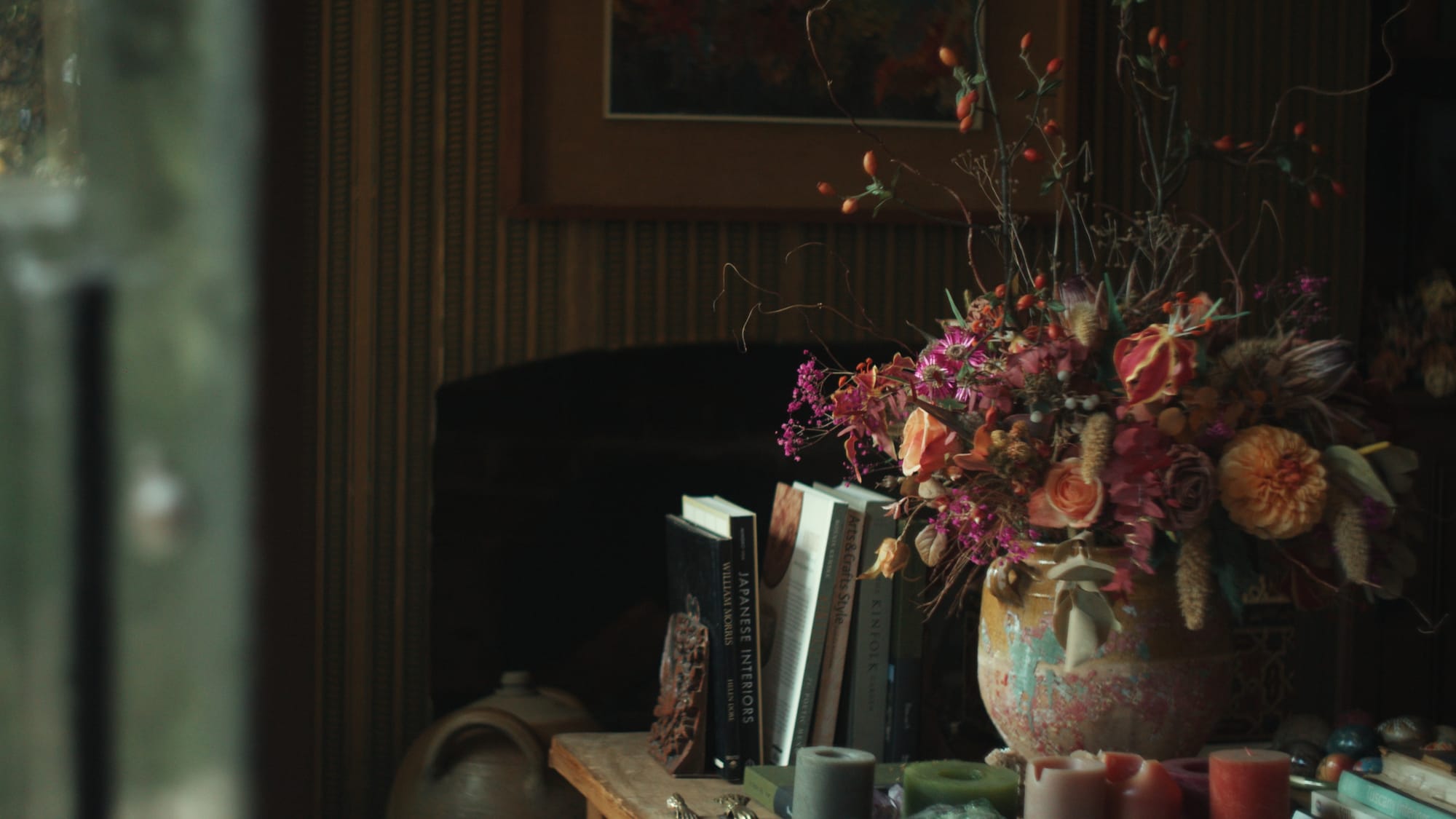 A wild, sculptural arrangement of dried flowers sits beside a stack of well-loved design books on a rustic wooden table
