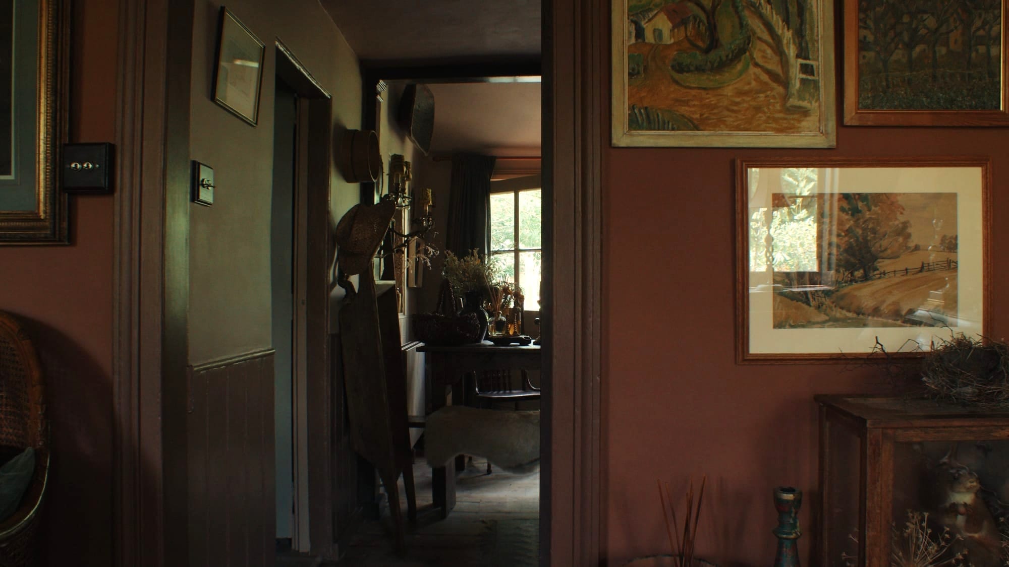 View through a doorway into a warm, rustic cottage interior with earthy walls, vintage paintings, dried flowers, and wooden furniture