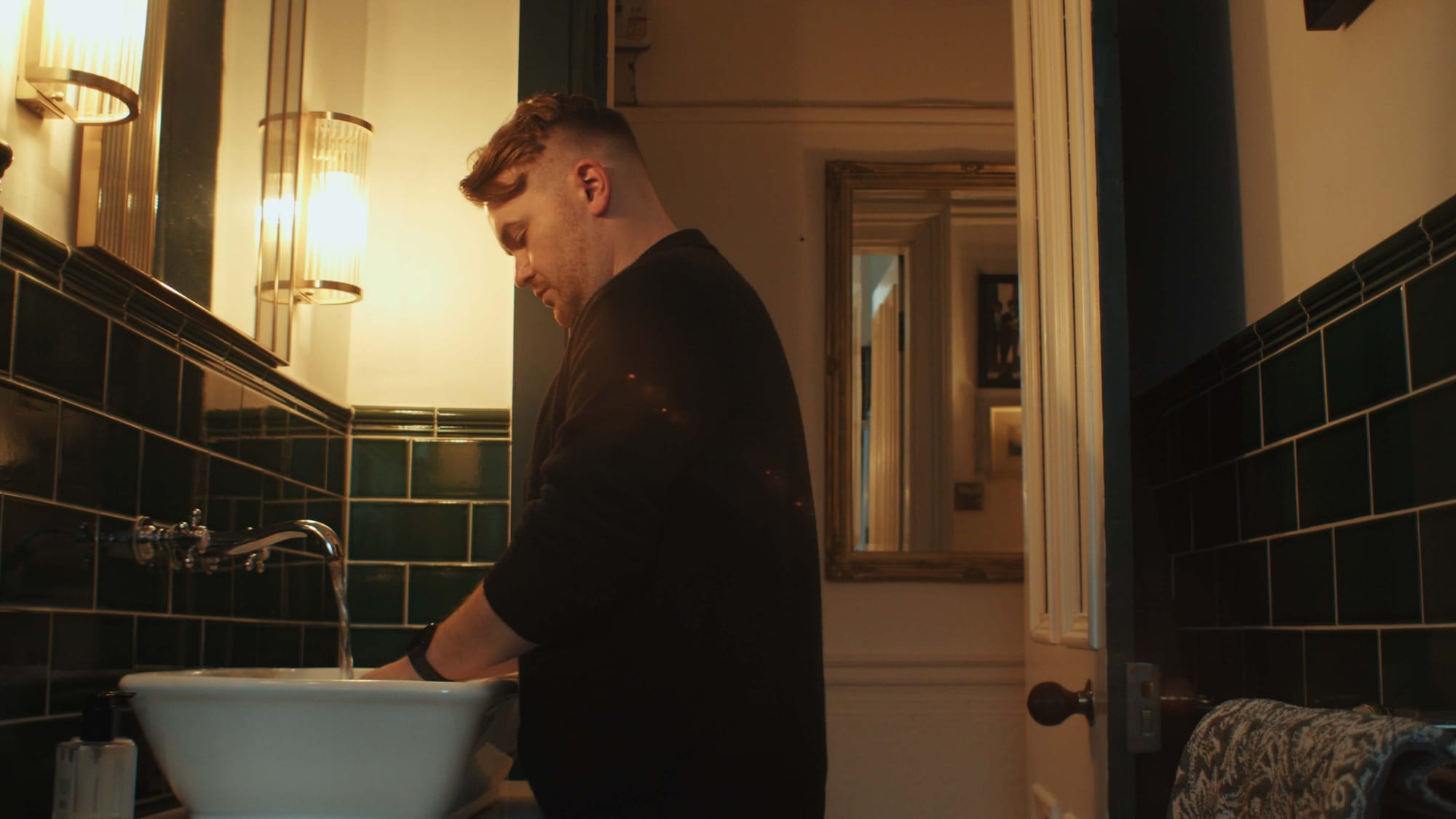 Man washing his hands in a moody bathroom with dark green tiles, warm lighting, and vintage details