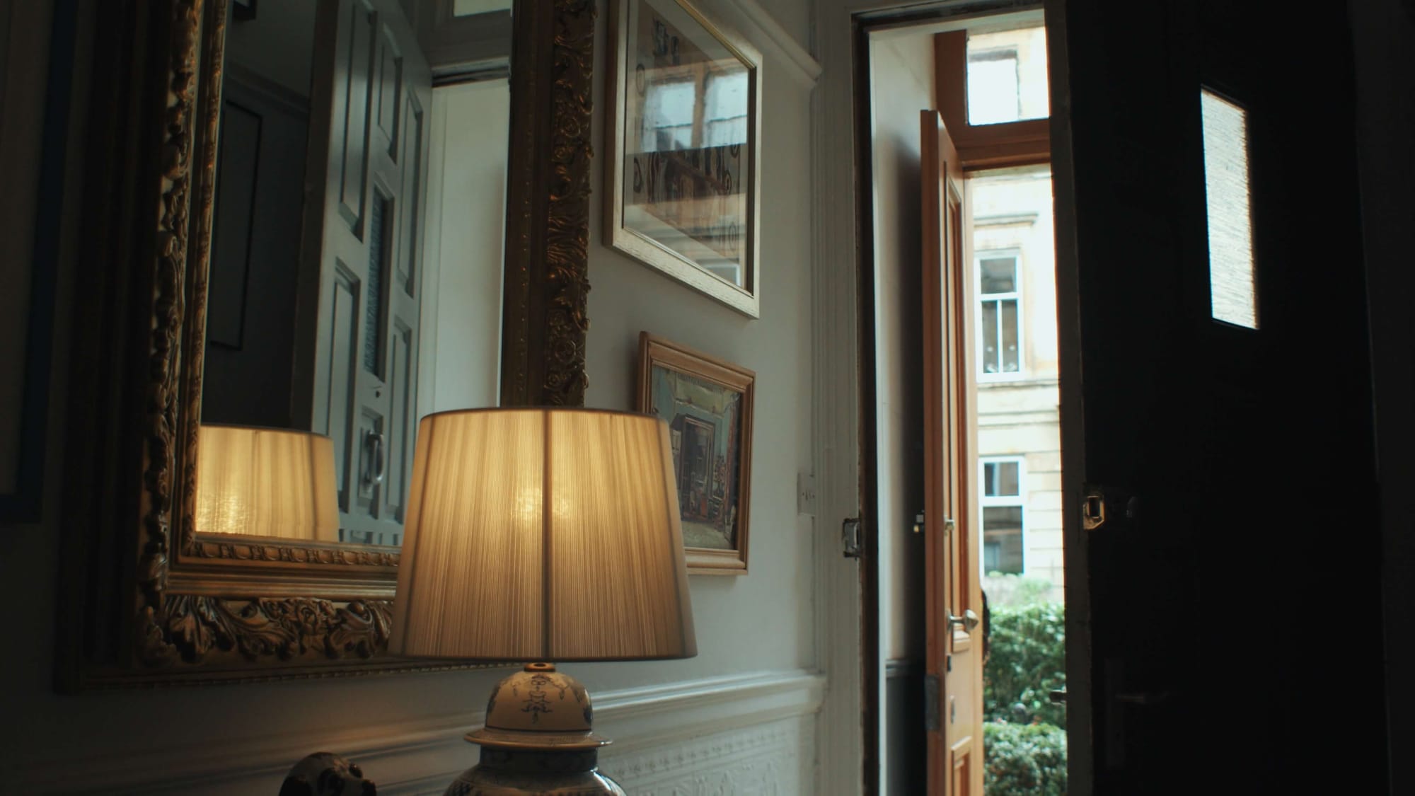 Bright hallway with an ornate mirror, framed art, and a glowing table lamp beside an open Victorian front door