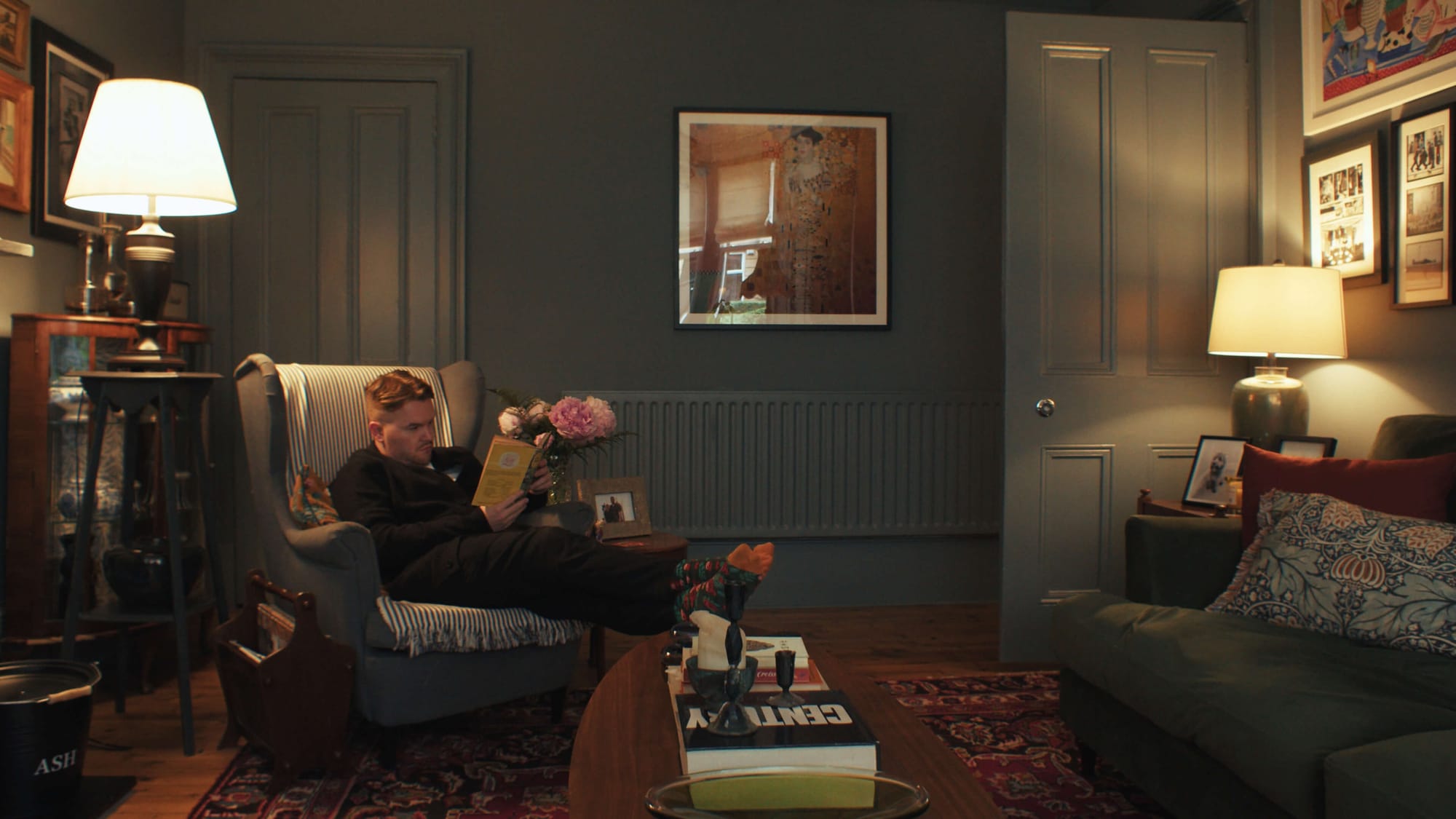 Cosy Victorian living room with a man reading in an armchair surrounded by warm lamps, artwork, and vintage décor