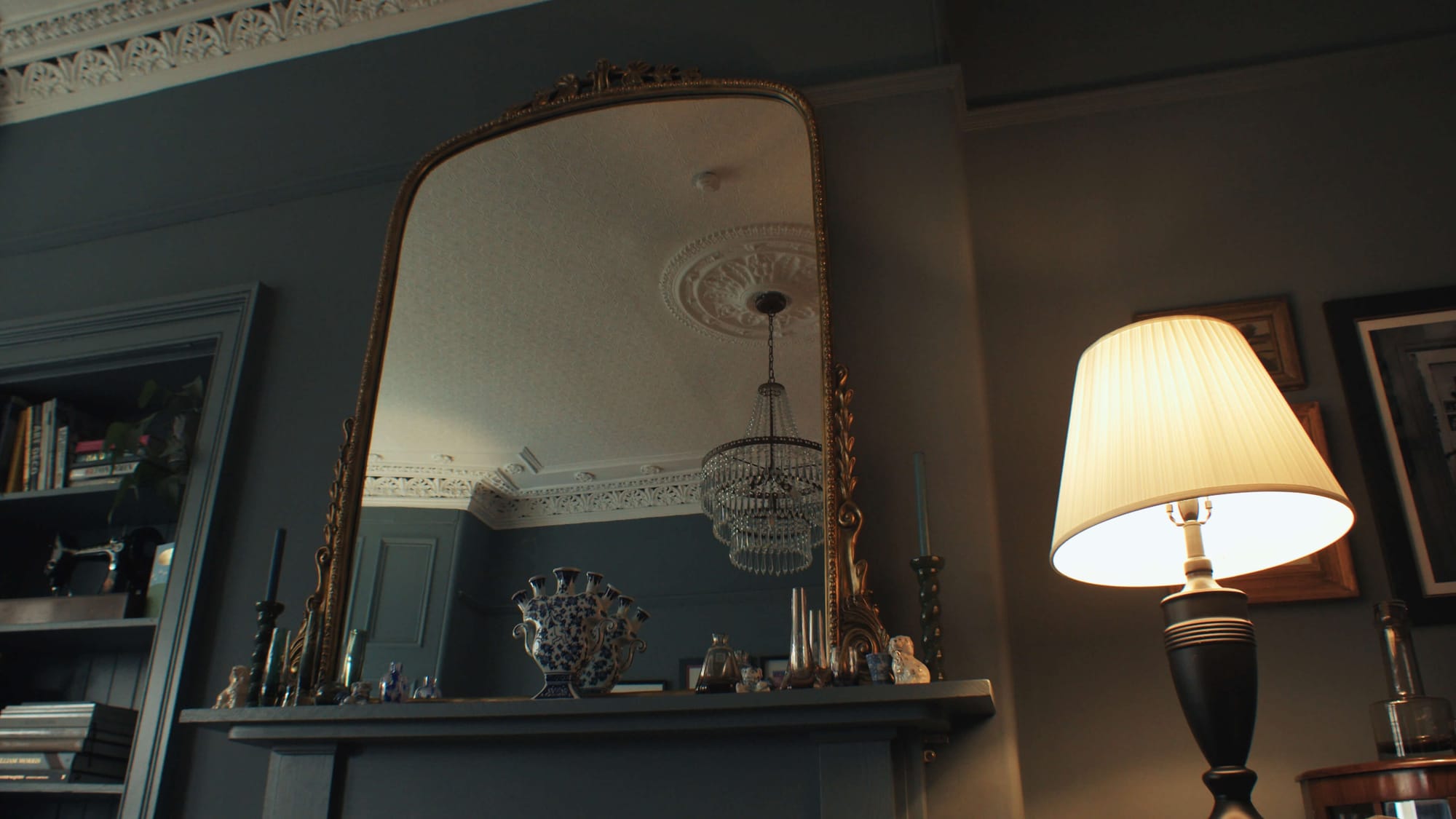 Ornate gold mirror above a mantel reflecting Victorian cornicing and a chandelier in a moody, characterful living room