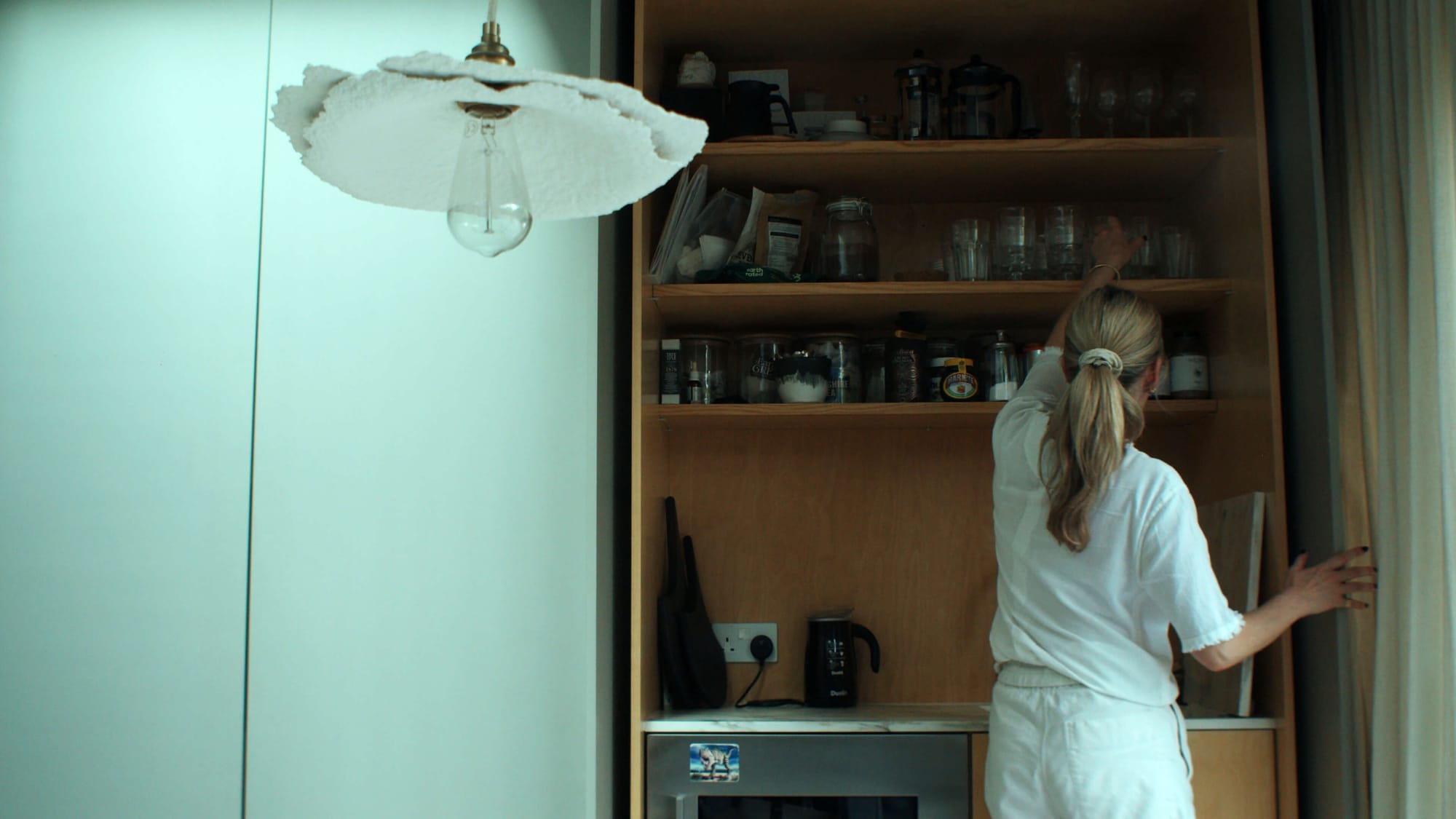 Louisa reaching for glassware on open wooden kitchen shelves beneath a hanging light