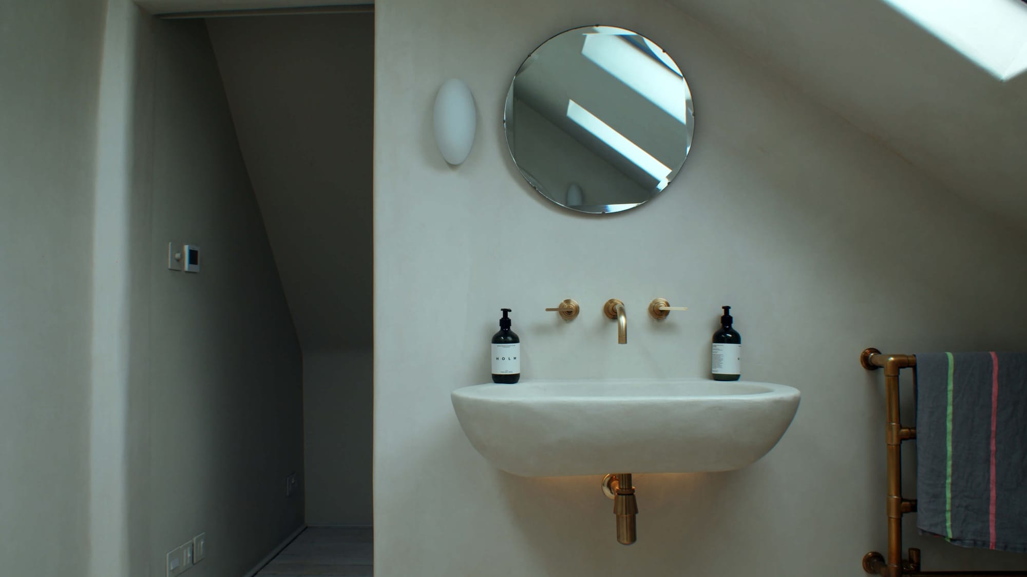 Wall-mounted white sink with brass tap and fittings, round mirror above, two dark soap bottles, and a brass towel rail with a striped towel in a softly lit bathroom