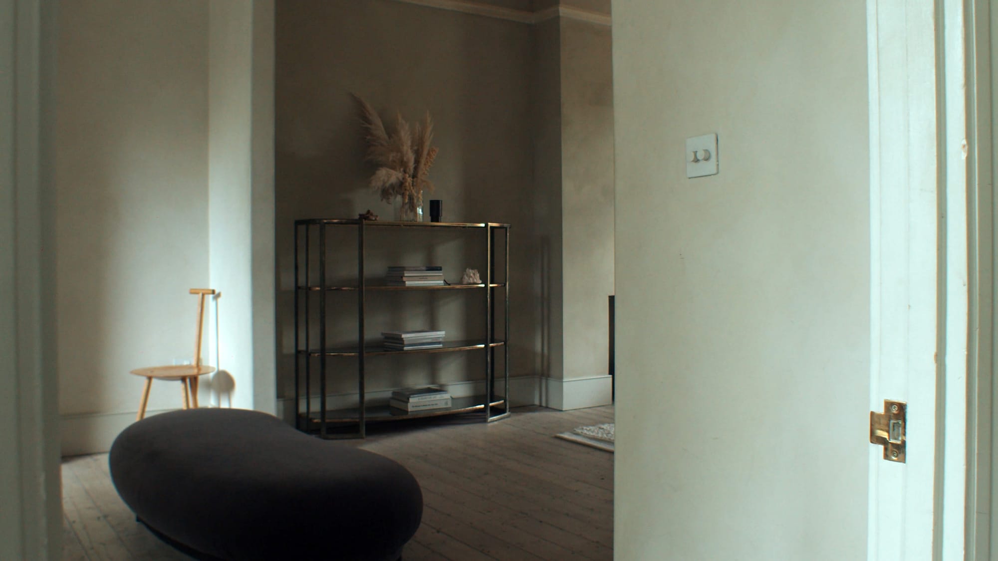 Minimal room with black shelving unit, stacked books, dried grasses in a vase, wooden chair and dark upholstered bench
